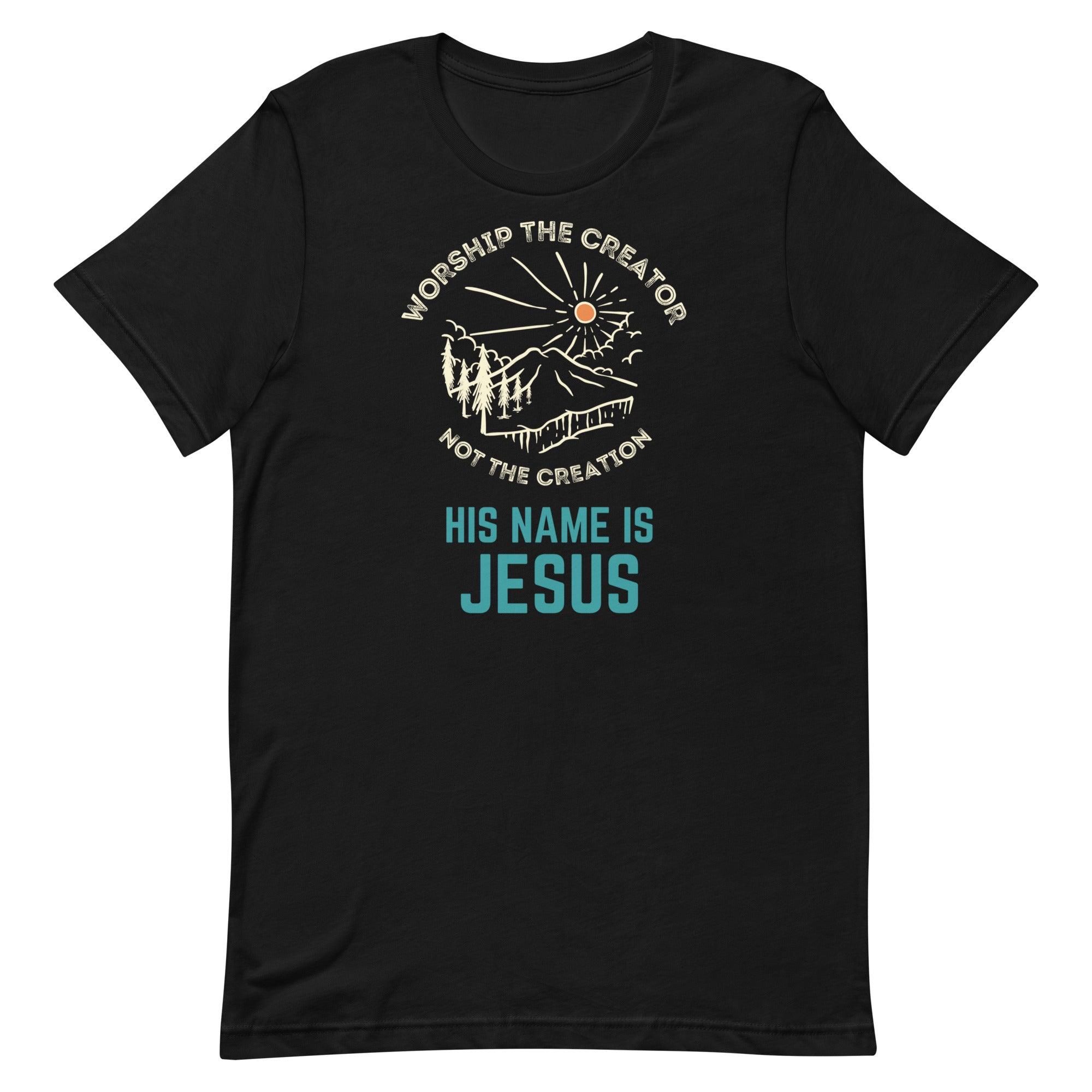 Flatlay of a black Christian t-shirt. There is a graphic of a mountain, trees, and sun. Over the graphic, it says "Worship the creator", and under it says "not the creation". On the bottom, it says "His name is Jesus". This shirt is sold at Amela&