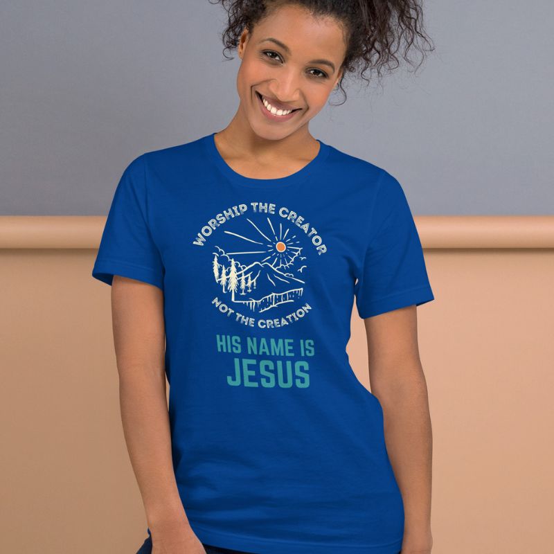 Black woman wearing a blue Christian t-shirt. There is a graphic of a mountain, trees, and sun. Over the graphic, it says &quot;Worship the creator&quot;, and under it says &quot;not the creation&quot;. On the bottom, it says &quot;His name is Jesus&quot;. This shirt is sold at Amela&
