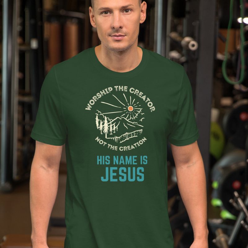 White man wearing a green Christian t-shirt. There is a graphic of a mountain, trees, and sun. Over the graphic, it says &quot;Worship the creator&quot;, and under it says &quot;not the creation&quot;. On the bottom, it says &quot;His name is Jesus&quot;. This shirt is sold at Amela&