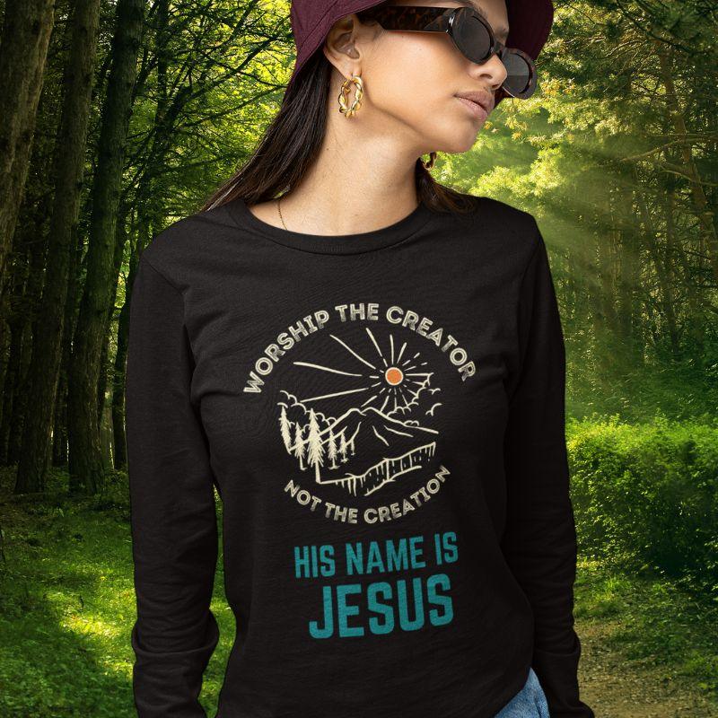 Asian woman wearing a black long sleeve Christian shirt. There is a graphic of a mountain, trees, and sun. Over the graphic, it says &quot;Worship the creator&quot;, and under it says &quot;not the creation&quot;. On the bottom, it says &quot;His name is Jesus&quot;. This shirt is sold at Amela&